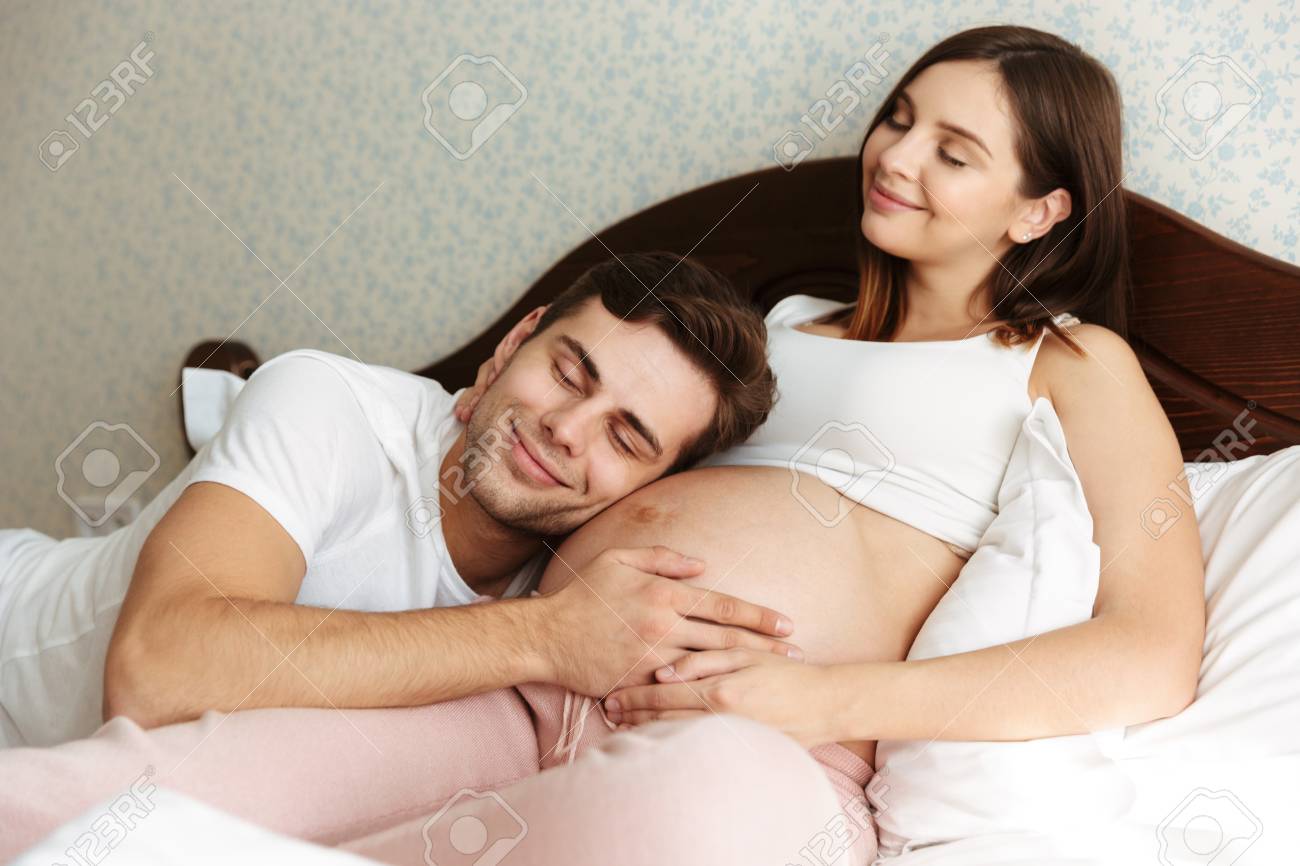 Delighted Young Pregnant Wife Lying In Bed With Her Husband In