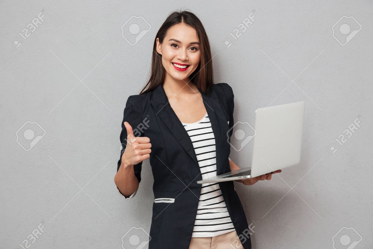 Holding asian business woman holding laptop computer and showing thumb up while looking at the camera over gray background - 93807278 Holding asian business woman holding laptop computer and showing thumb up while looking at the camera over gray background - 93807278