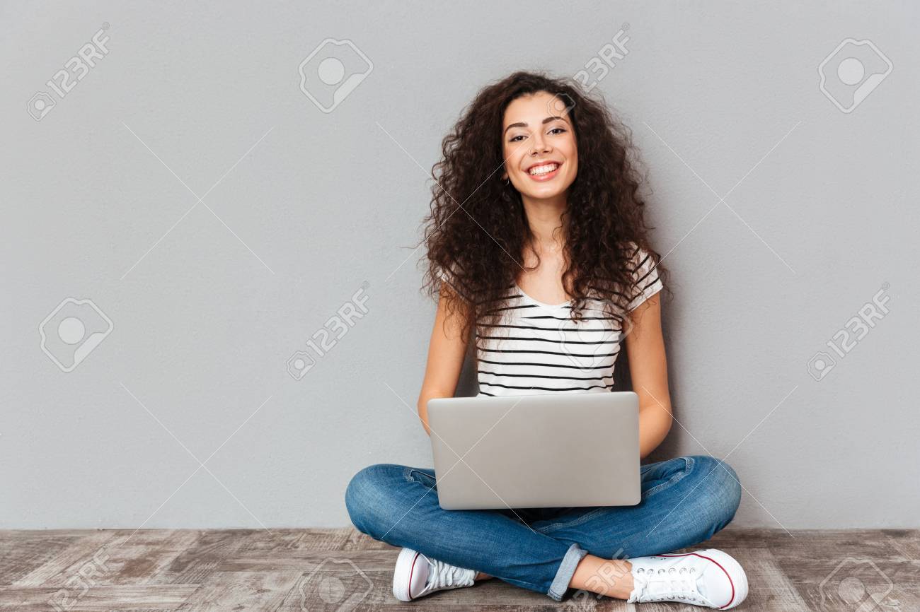 Portrait of satisfied female with beautiful smile enjoying watching movie in silver computer and sitting in lotus pose on the floor over grey wall - 94077242 Portrait of satisfied female with beautiful smile enjoying watching movie in silver computer and sitting in lotus pose on the floor over grey wall - 94077242