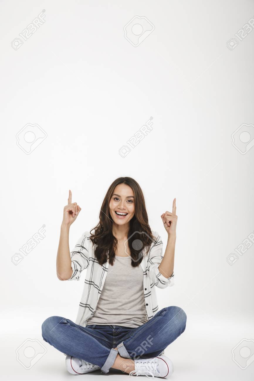 床に蓮のポーズをとって 白い壁の上に隔離された人差し指を上に向けるカジュアルな服を着た喜んだ女性のフルレングスの肖像画 の写真素材 画像素材 Image