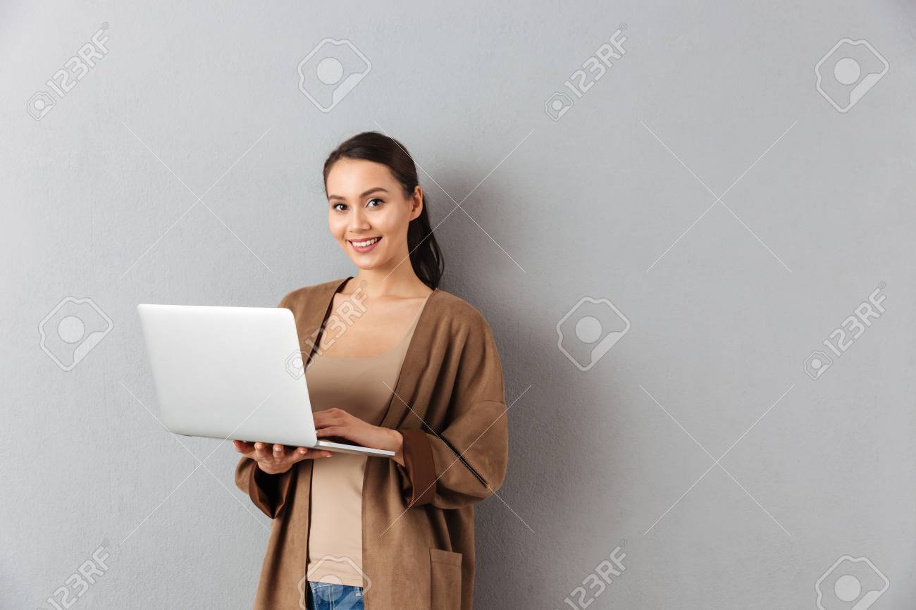 Portrait of a happy young asian woman holding laptop computer while standing and looking at camera over gray background - 95133518 Portrait of a happy young asian woman holding laptop computer while standing and looking at camera over gray background - 95133518