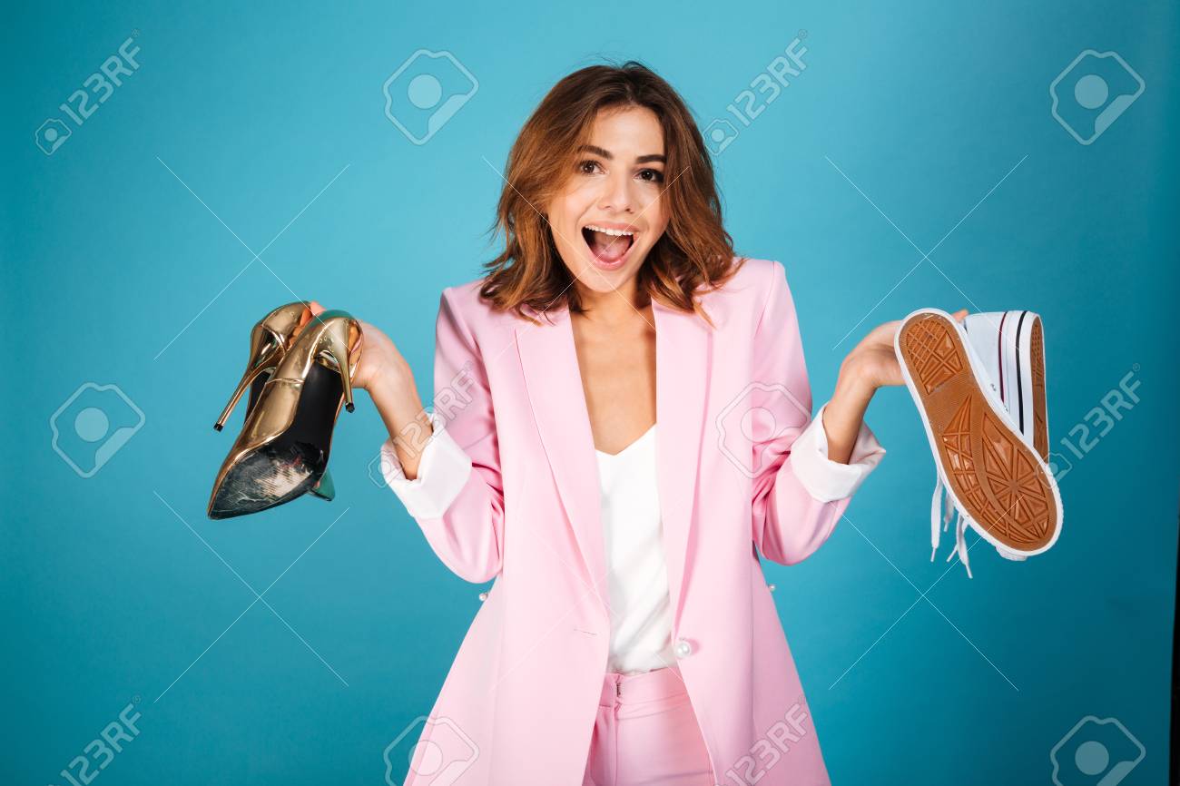 ピンクのスーツに身を包んだ陽気な女性の肖像画は ハイヒールの靴と青い背景の上に隔離されたスニーカーのペアを保持しています の写真素材 画像素材 Image