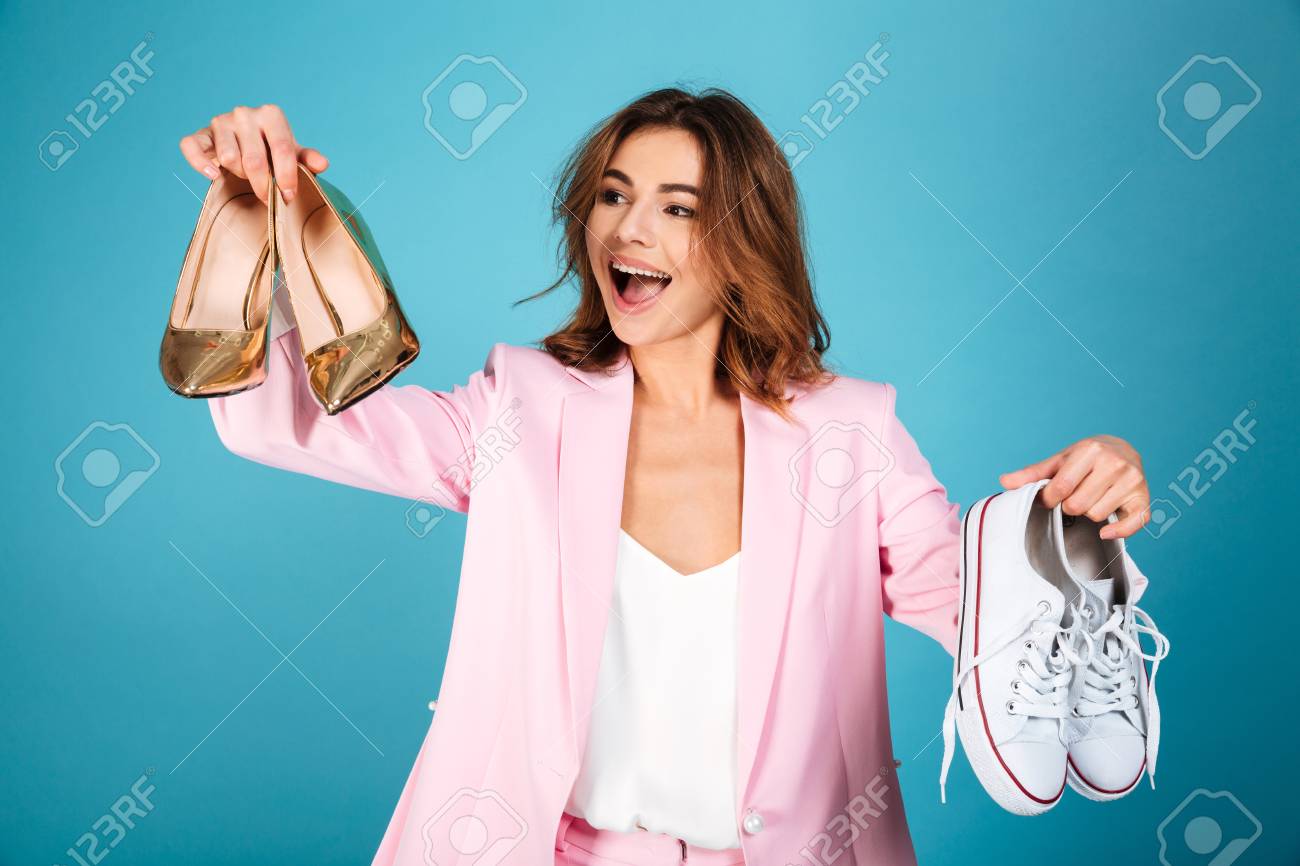 ピンクのスーツに身を包んだ幸せな女性の肖像画は ハイヒールの靴と青い背景の上に隔離されたスニーカーのペアを保持しています の写真素材 画像素材 Image