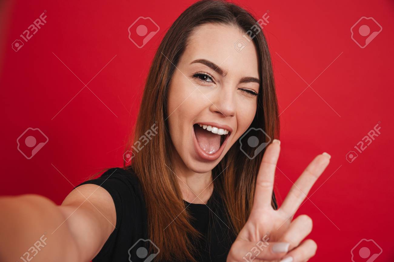 Cute woman 20s in black t-shirt having fun and taking selfie with gesturing peace sign isolated over red background - 97800706 Cute woman 20s in black t-shirt having fun and taking selfie with gesturing peace sign isolated over red background - 97800706
