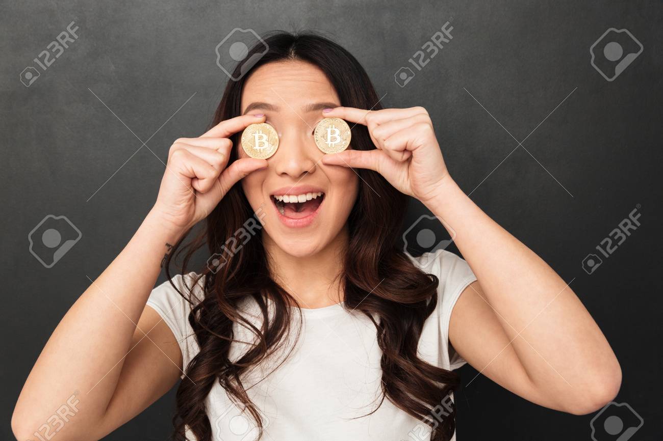 目を覆う2つの黄金のビットコインを保持している黒い壁の背景の上に孤立して立っている興奮した感情的なアジアの若い女性のイメージ。の写真素材・画像素材  Image 97983322