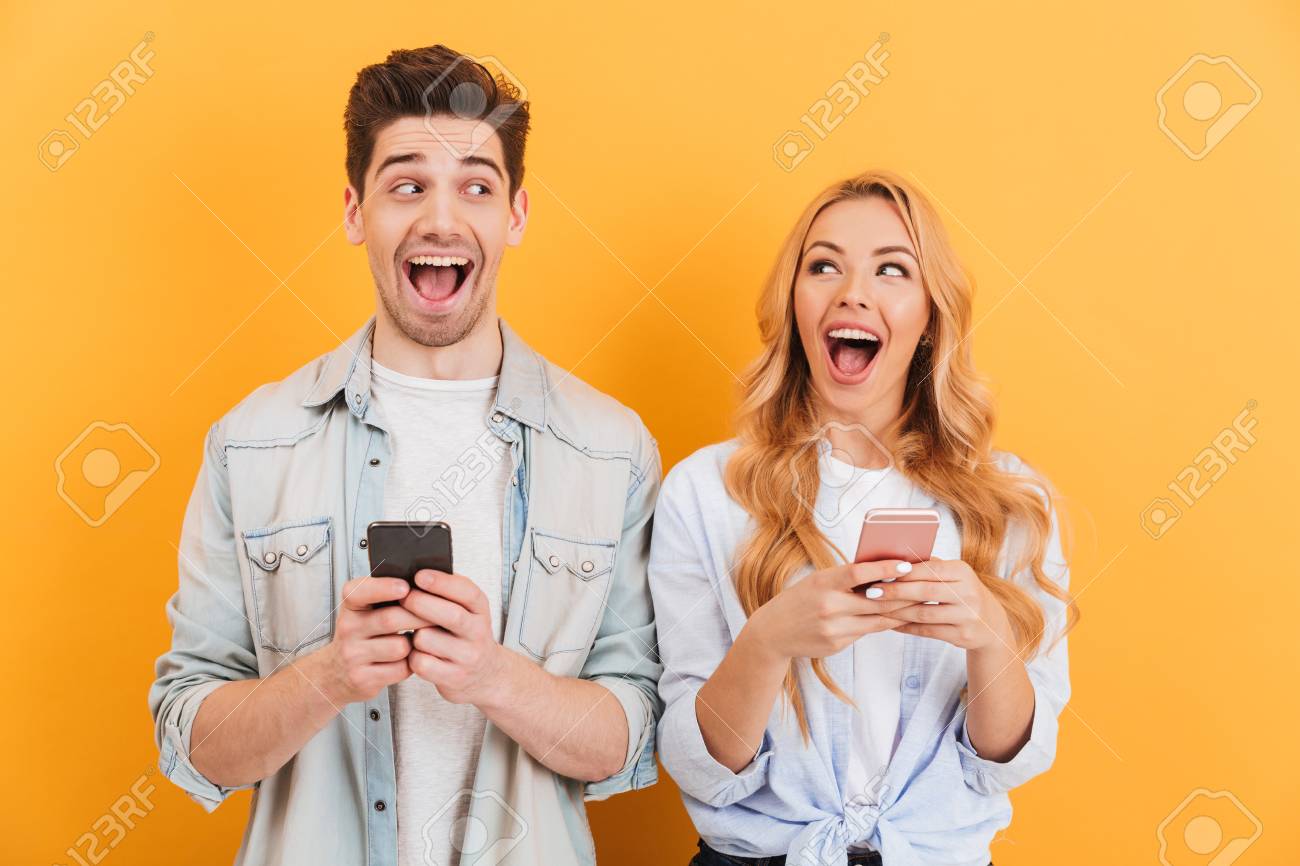 Photo of cheerful man and woman looking at each other with happy emotions while using smartphones isolated over yellow background - 103929568 Photo of cheerful man and woman looking at each other with happy emotions while using smartphones isolated over yellow background - 103929568