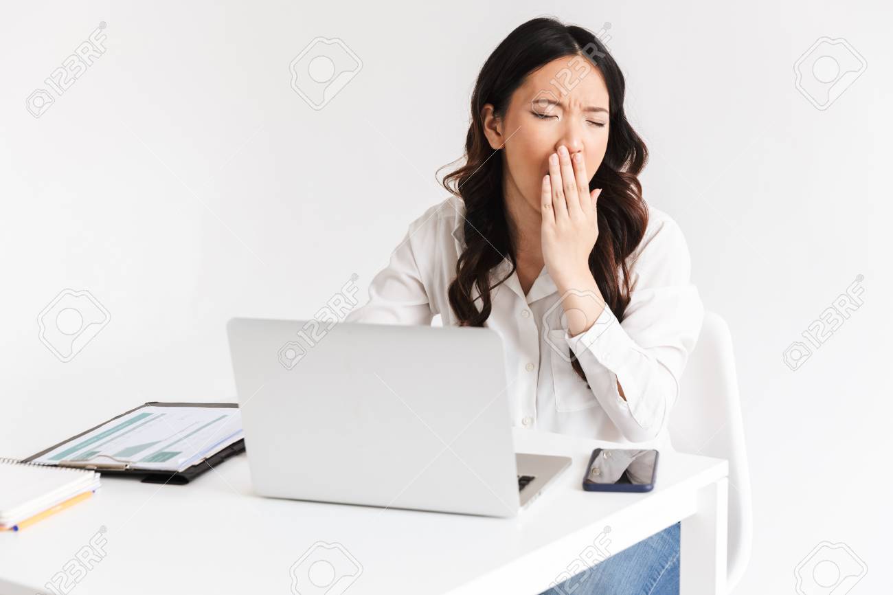 Photo of overworked tired asian business woman 20s wearing office clothing yawning while sitting at table with laptop open isolated over white background - 107728955 Photo of overworked tired asian business woman 20s wearing office clothing yawning while sitting at table with laptop open isolated over white background - 107728955