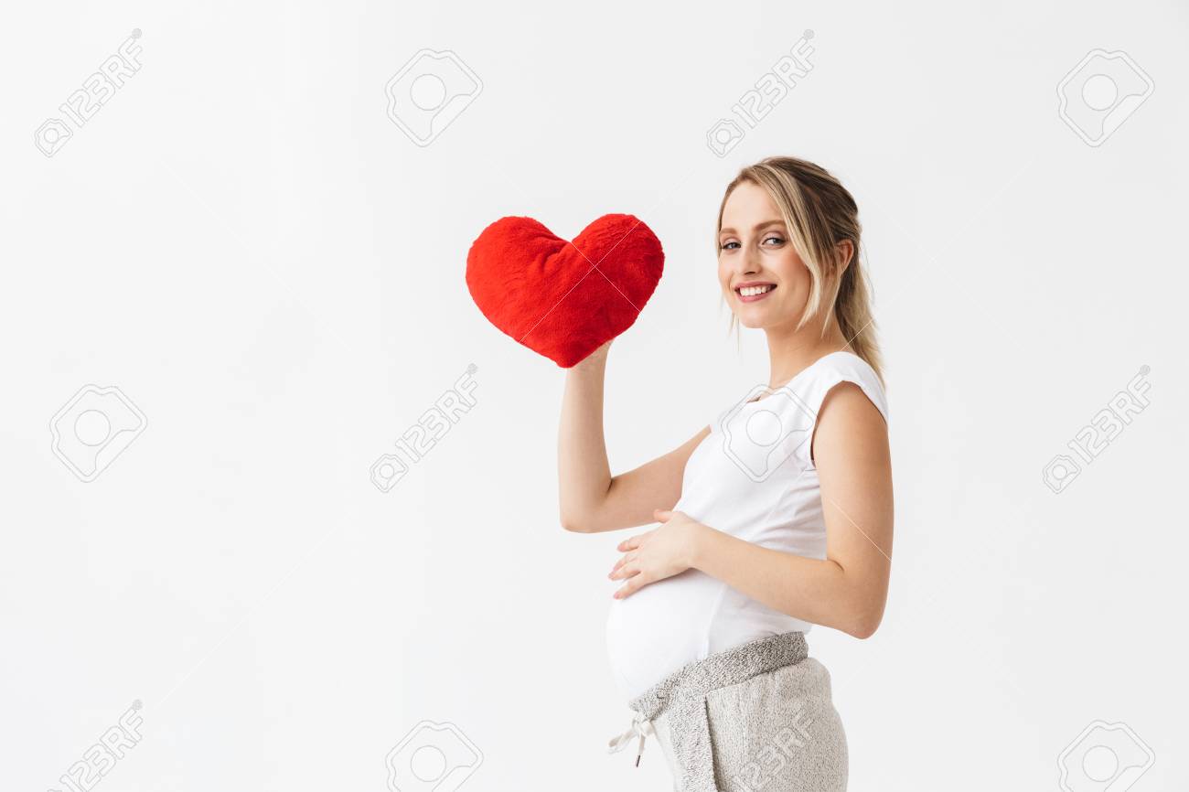 Beautiful young pregnant woman standing isolated over white background, holding heart - 120563976 Beautiful young pregnant woman standing isolated over white background, holding heart - 120563976