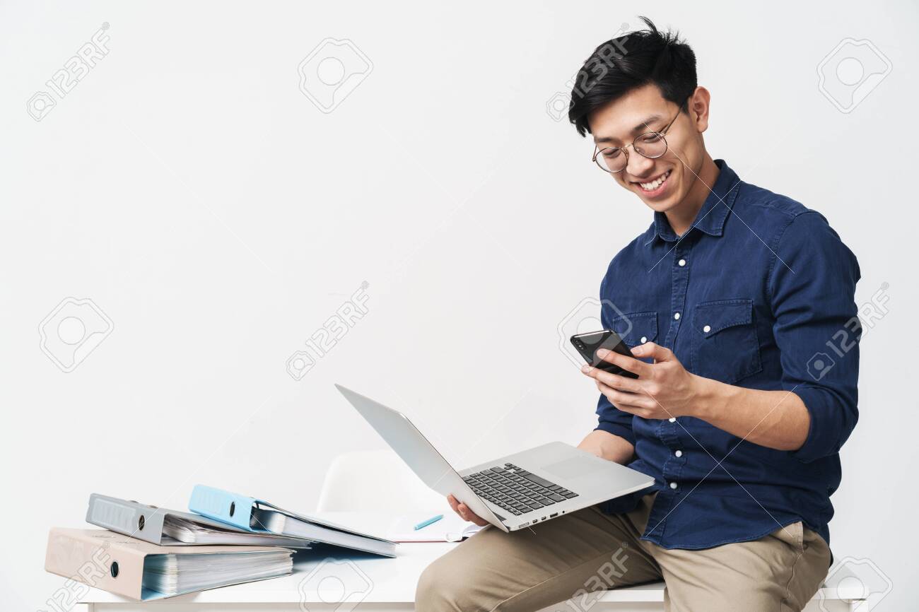 Photo of smiling asian man 20s wearing eyeglasses holding smartphone and laptop while working in office isolated over white background - 122915556 Photo of smiling asian man 20s wearing eyeglasses holding smartphone and laptop while working in office isolated over white background - 122915556