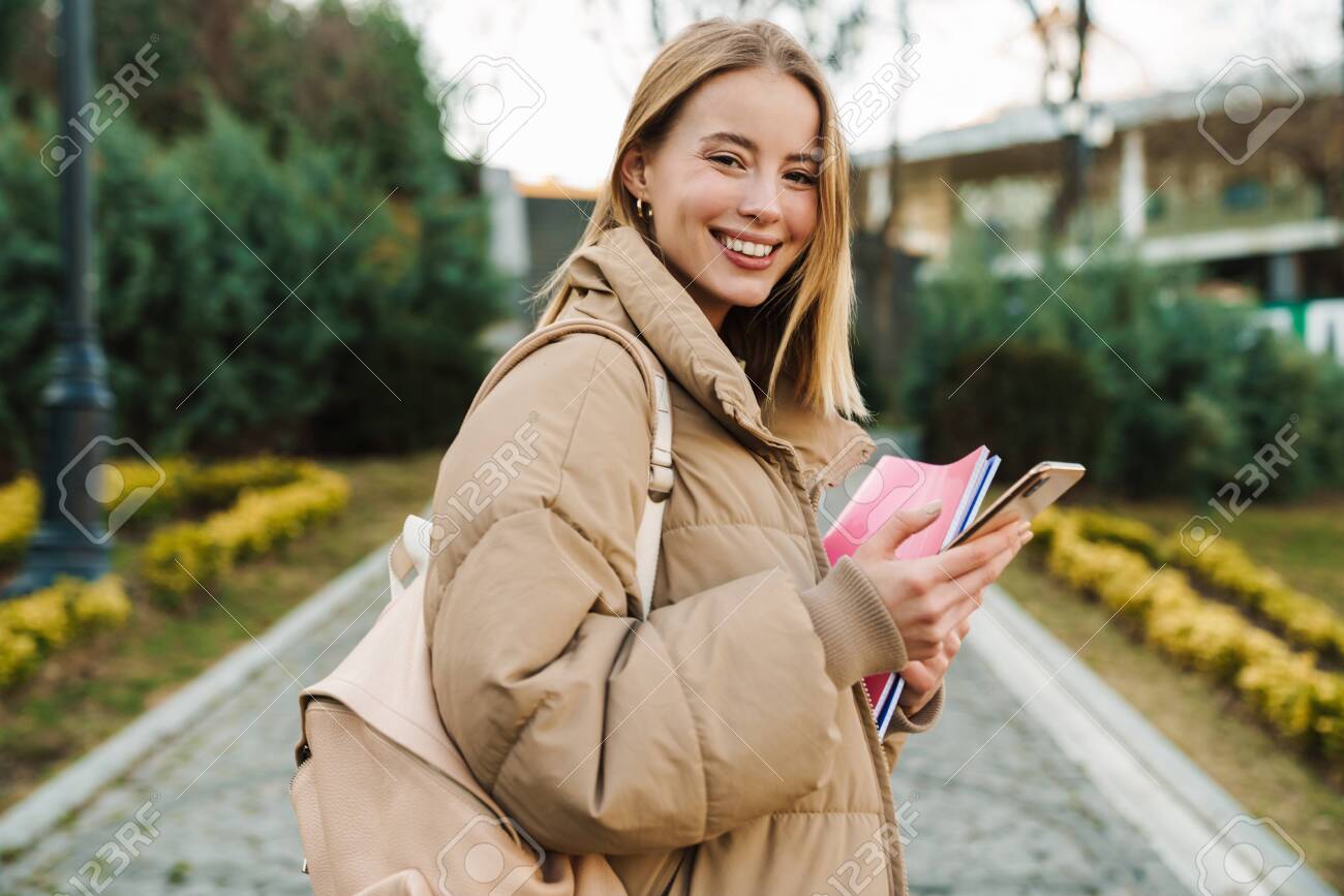 Portrait of smiling young woman holding exercise books and using mobile phone while walking in park - 148360425 Portrait of smiling young woman holding exercise books and using mobile phone while walking in park - 148360425
