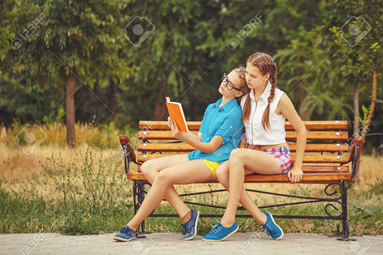 Las Mejores Amigas Están Leyendo Un Libro Mientras Está Sentado En Un En Parque De Verano. La Muchacha Adolescente Vestido Con Pantalones Cortos Y Una Camisa. En Las Vacaciones De