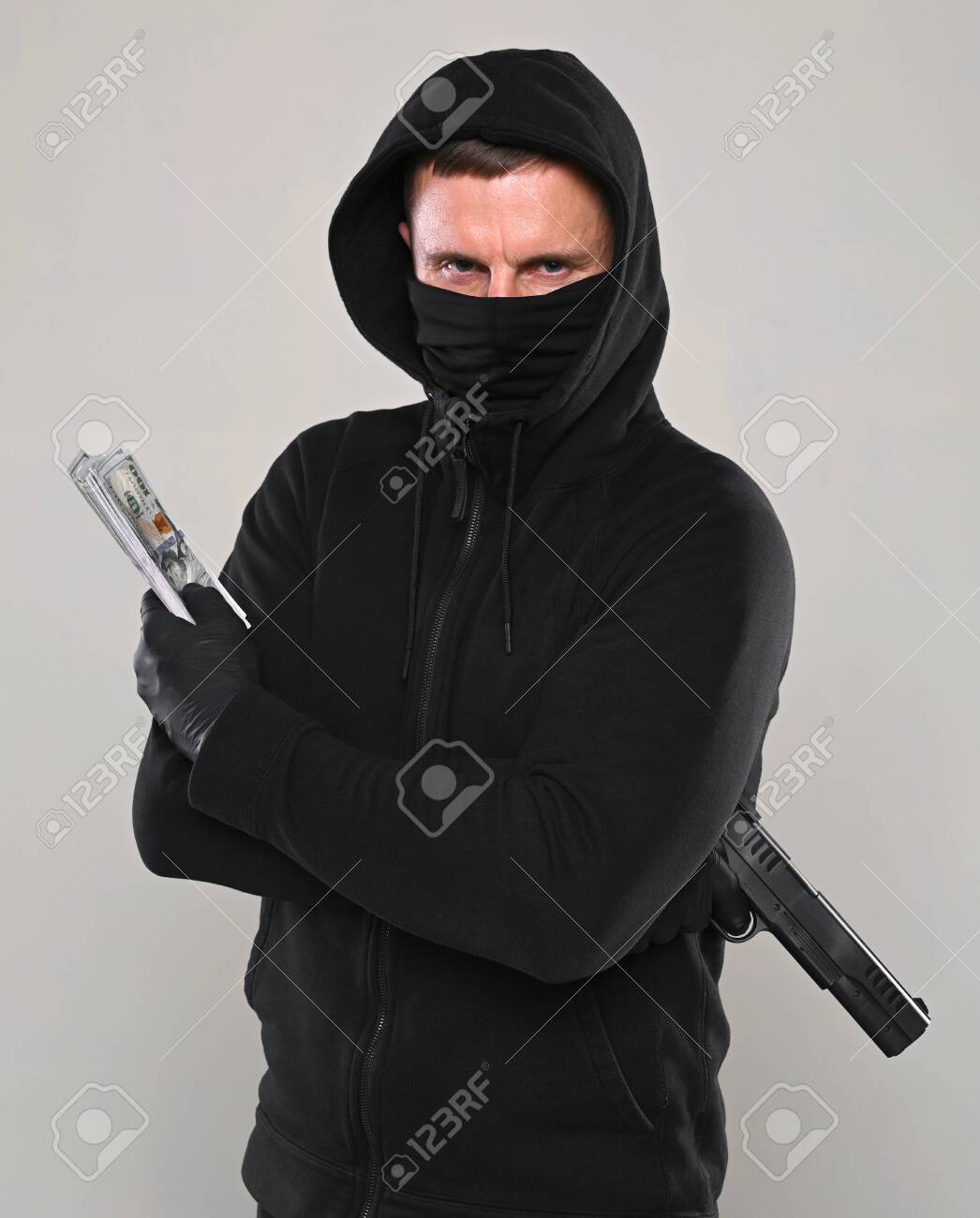 Man in black mask and hoody with a gun and pack of dollars over gray background - 143163093 Man in black mask and hoody with a gun and pack of dollars over gray background - 143163093