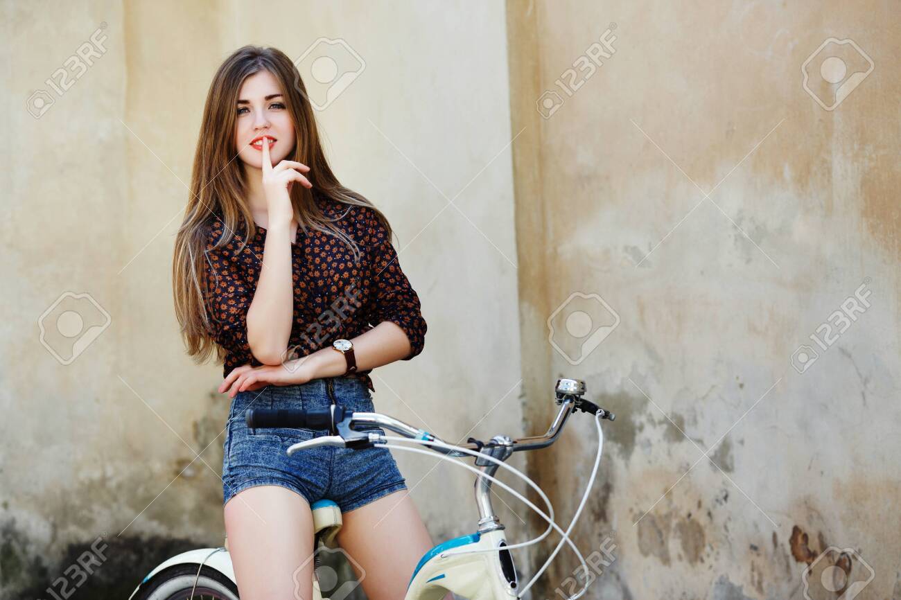 Smiling young woman with long hair is posing on the bicycle on the old wall background - 122488804 Smiling young woman with long hair is posing on the bicycle on the old wall background - 122488804