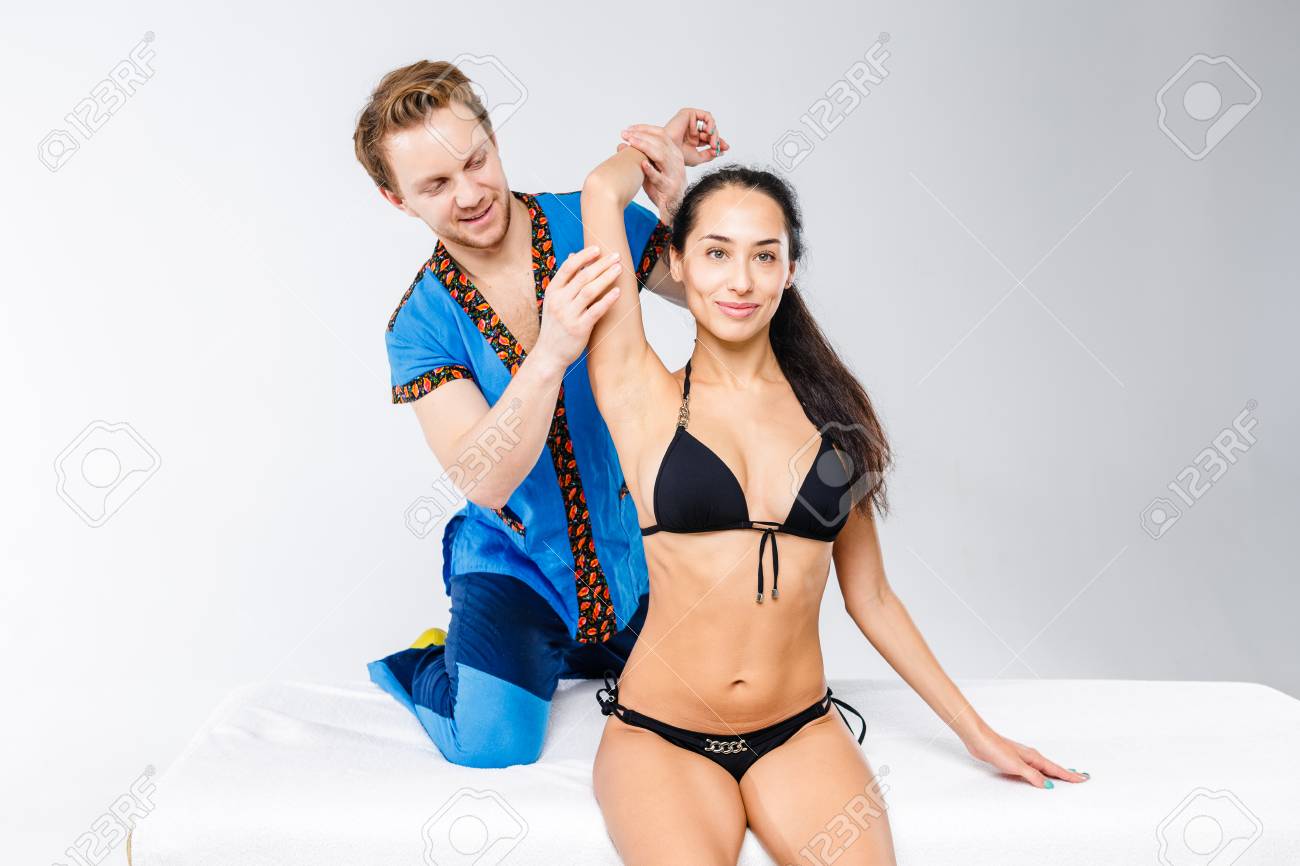 テーママッサージとボディケア 作業スーツの若い医師のマッサージ師は 黒い水着でテーブルの上に座っている美しい女の子の首 肩 手の筋肉を伸ばする ためのマッサージを行います の写真素材 画像素材 Image