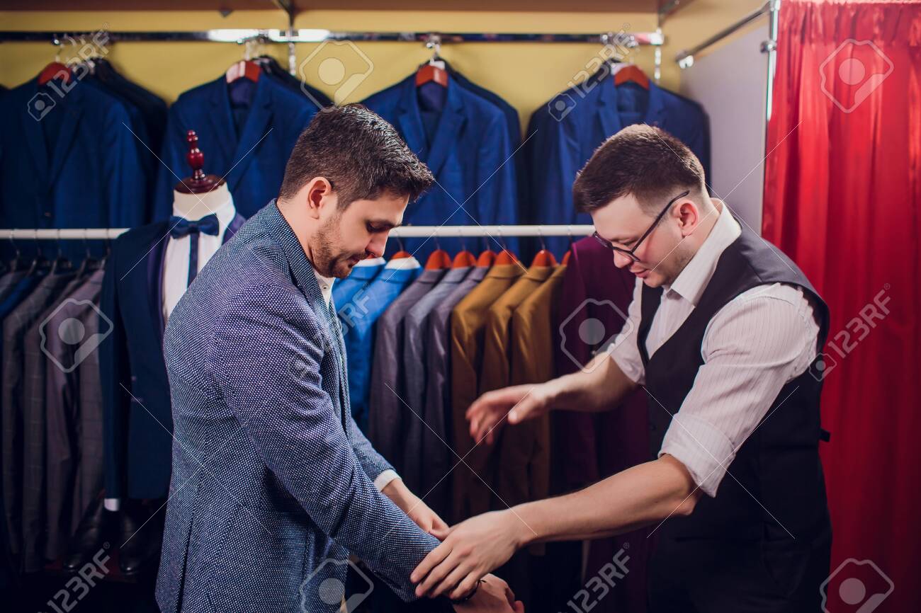 Businessman in classic vest against row suits in shop. Man helps another try on suit in clothing store - 121333141 Businessman in classic vest against row suits in shop. Man helps another try on suit in clothing store - 121333141