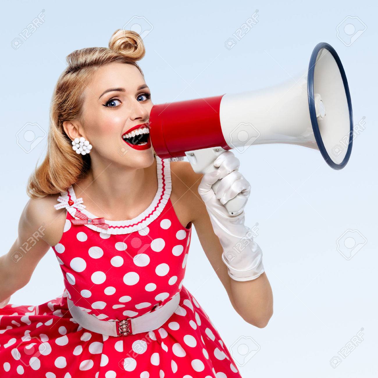 Portrait of woman holding megaphone, dressed in pin-up style red dress in polka dot and white gloves, on blue background. Caucasian blond model posing in retro fashion vintage studio shoot. - 65643469 Portrait of woman holding megaphone, dressed in pin-up style red dress in polka dot and white gloves, on blue background. Caucasian blond model posing in retro fashion vintage studio shoot. - 65643469