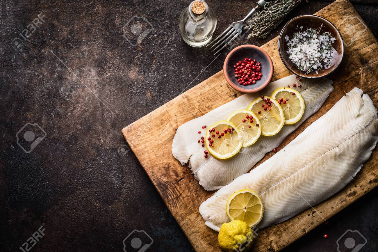 Raw cod fish fillet with lemon slices and herbal salt on rustic wooden cutting board on dark background, top view. Fish cooking preparation. Healthy diet food. Border. Copy space - 128752553 Raw cod fish fillet with lemon slices and herbal salt on rustic wooden cutting board on dark background, top view. Fish cooking preparation. Healthy diet food. Border. Copy space - 128752553