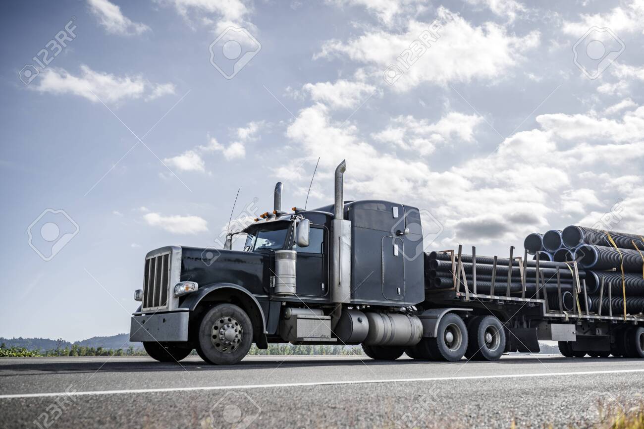 Big rig powerful professional industrial bonnet semi truck for long haul delivery commercial plastic pipes going with step down semi trailer on the summer road with green forest trees on the sides - 129624280 Big rig powerful professional industrial bonnet semi truck for long haul delivery commercial plastic pipes going with step down semi trailer on the summer road with green forest trees on the sides - 129624280