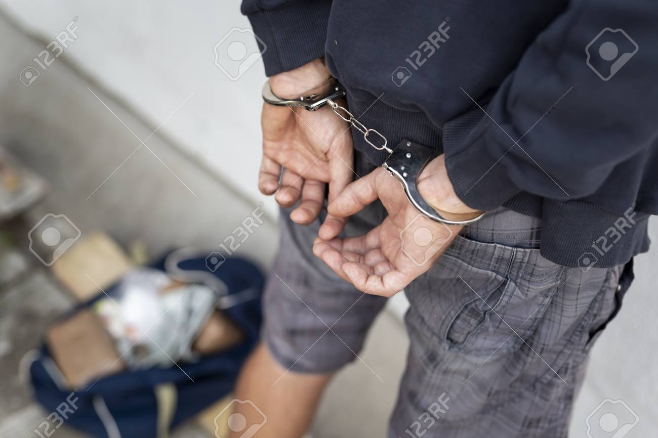 Drug dealer under arrest confined with handcuffs and hands at his back, standing next to a wall. Focus on the handcuffs chain - 108204877 Drug dealer under arrest confined with handcuffs and hands at his back, standing next to a wall. Focus on the handcuffs chain - 108204877