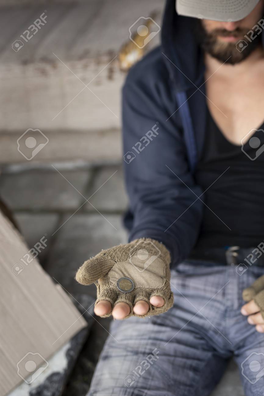 キャップと指なし手袋の上にパーカーを着て、通りに座って、お金を物乞いしているホームレスの男性。手の中のコインに焦点を当てるの写真素材・画像素材  Image 111657971
