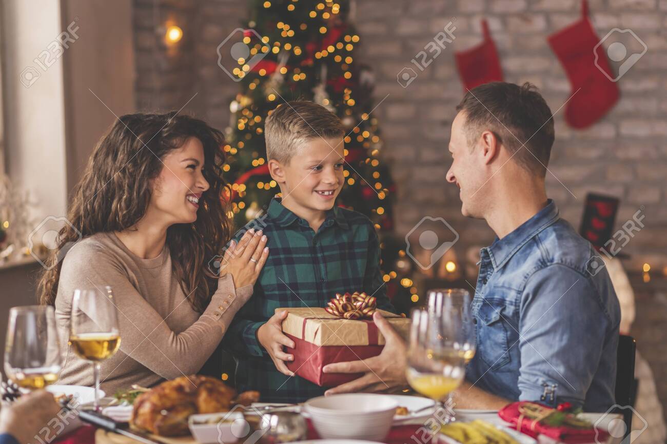Family having Christmas dinner at home, gathered around the table, enjoying their time together; mother and father giving Christmas present to their son - 158283320 Family having Christmas dinner at home, gathered around the table, enjoying their time together; mother and father giving Christmas present to their son - 158283320