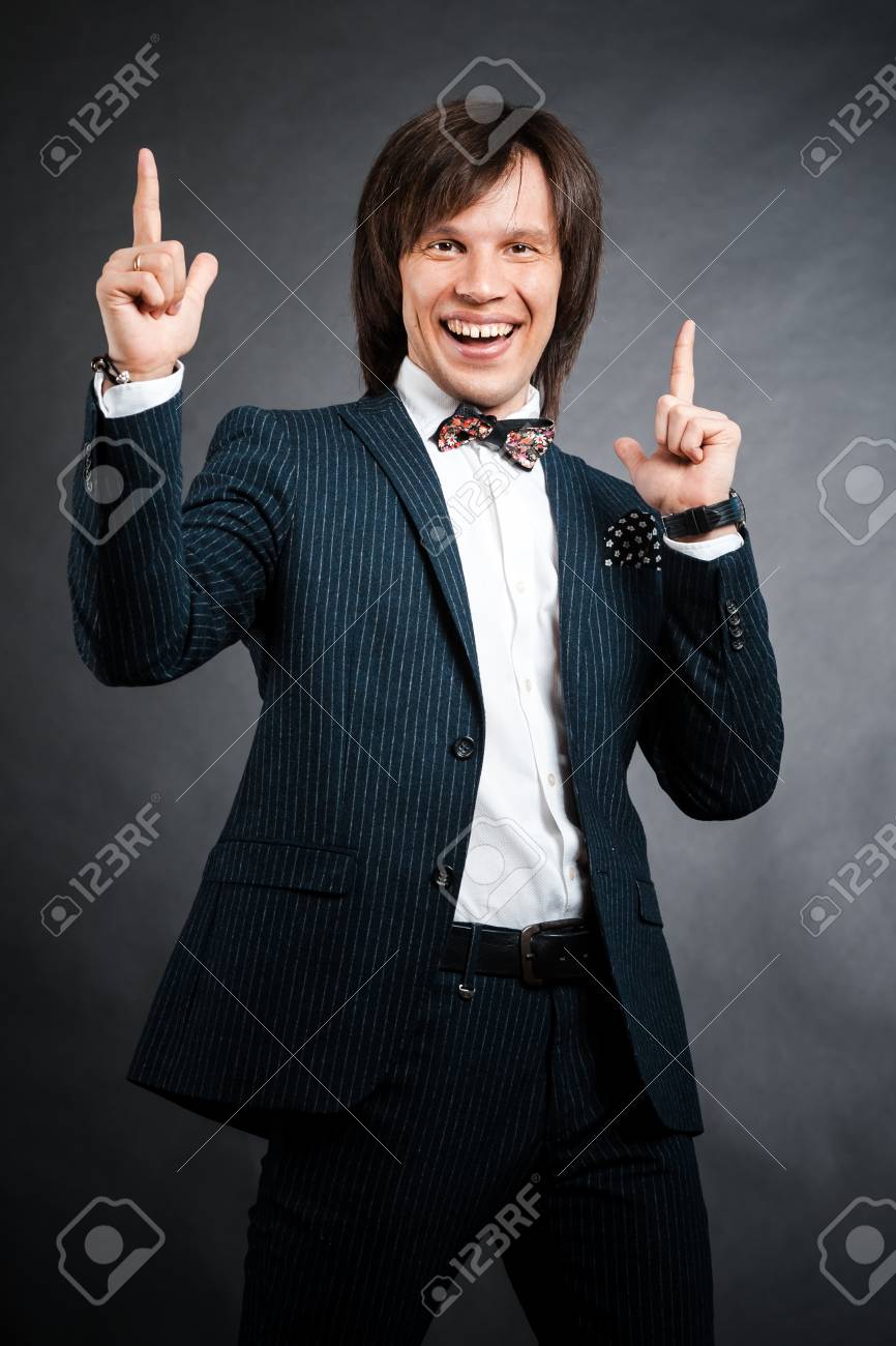 長い髪ブルネットと茶色の目でリクルート スーツ ストライプ蝶ネクタイと黒の背景で楽しみを示す 2 本の指で幸せなハンサムな男 の写真素材 画像素材 Image