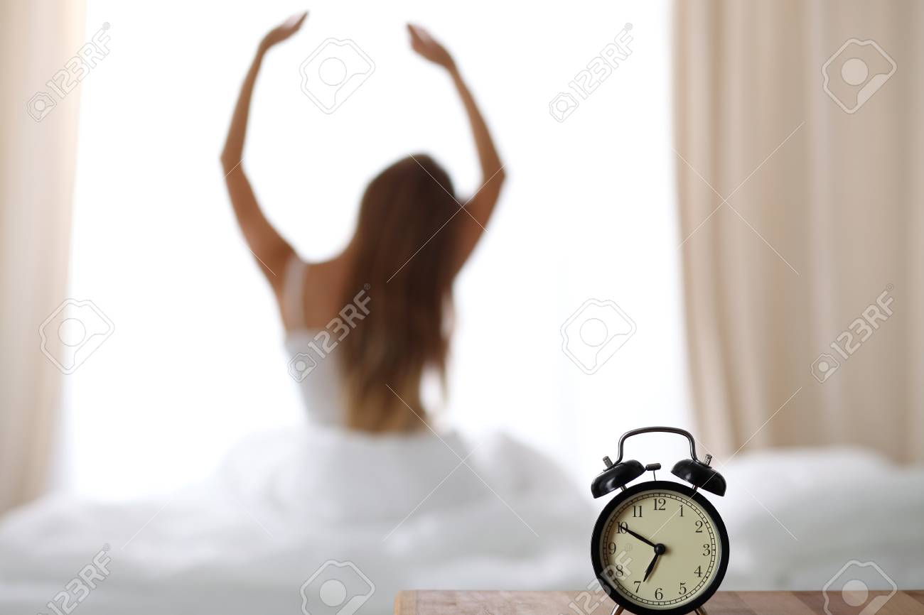 Réveil debout sur la table de chevet a déjà sonné tôt le matin pour se réveiller la femme est étirement dans son lit en arrière plan. Éveil précoce, Réveil debout sur la table de chevet a déjà sonné tôt le matin pour se réveiller la femme est étirement dans son lit en arrière plan. Éveil précoce,