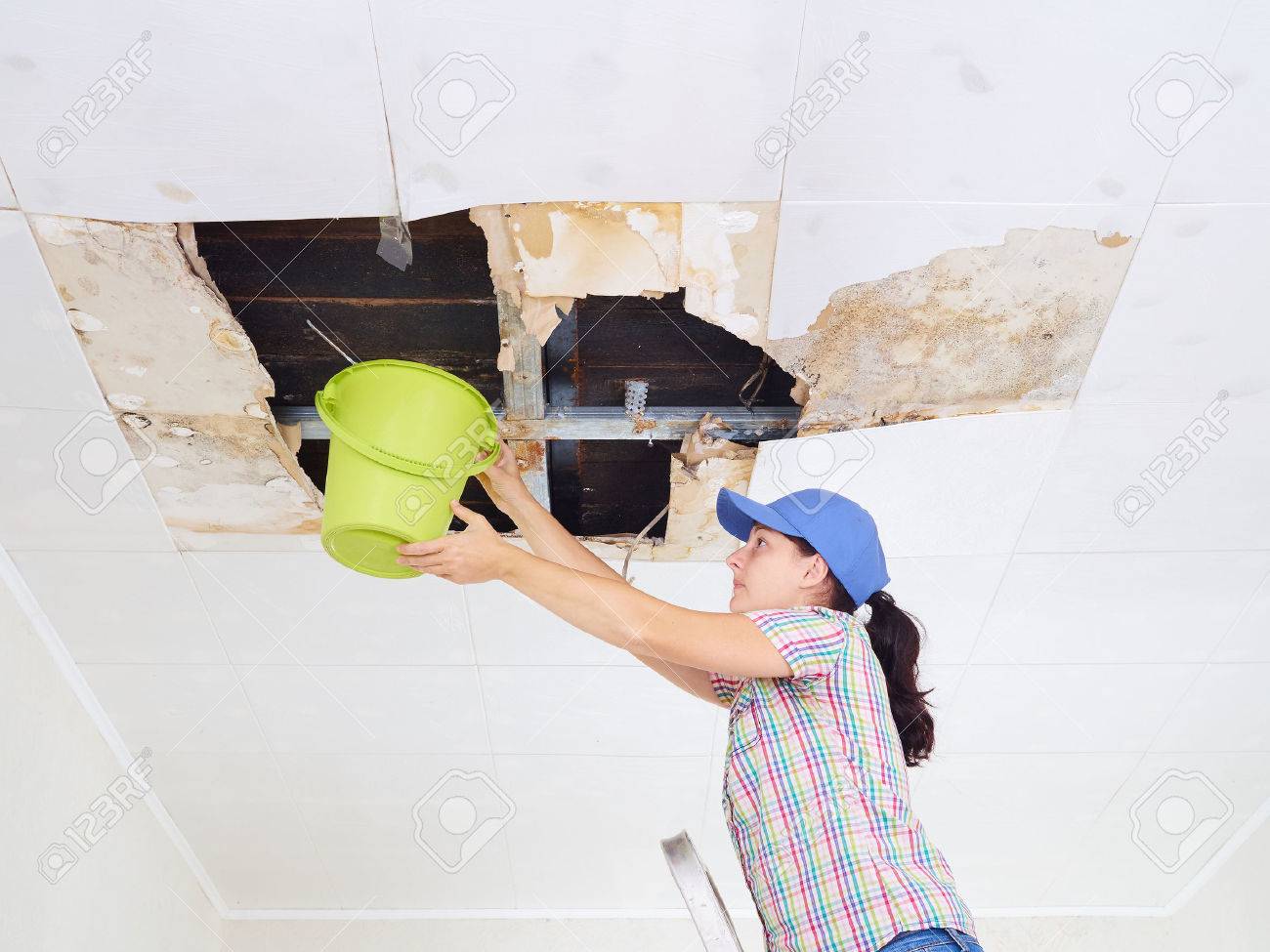 Young Woman Collecting Water In Bucket From Ceiling. Ceiling panels damaged huge hole in roof from rainwater leakage.Water damaged ceiling . - 66447462 Young Woman Collecting Water In Bucket From Ceiling. Ceiling panels damaged huge hole in roof from rainwater leakage.Water damaged ceiling . - 66447462