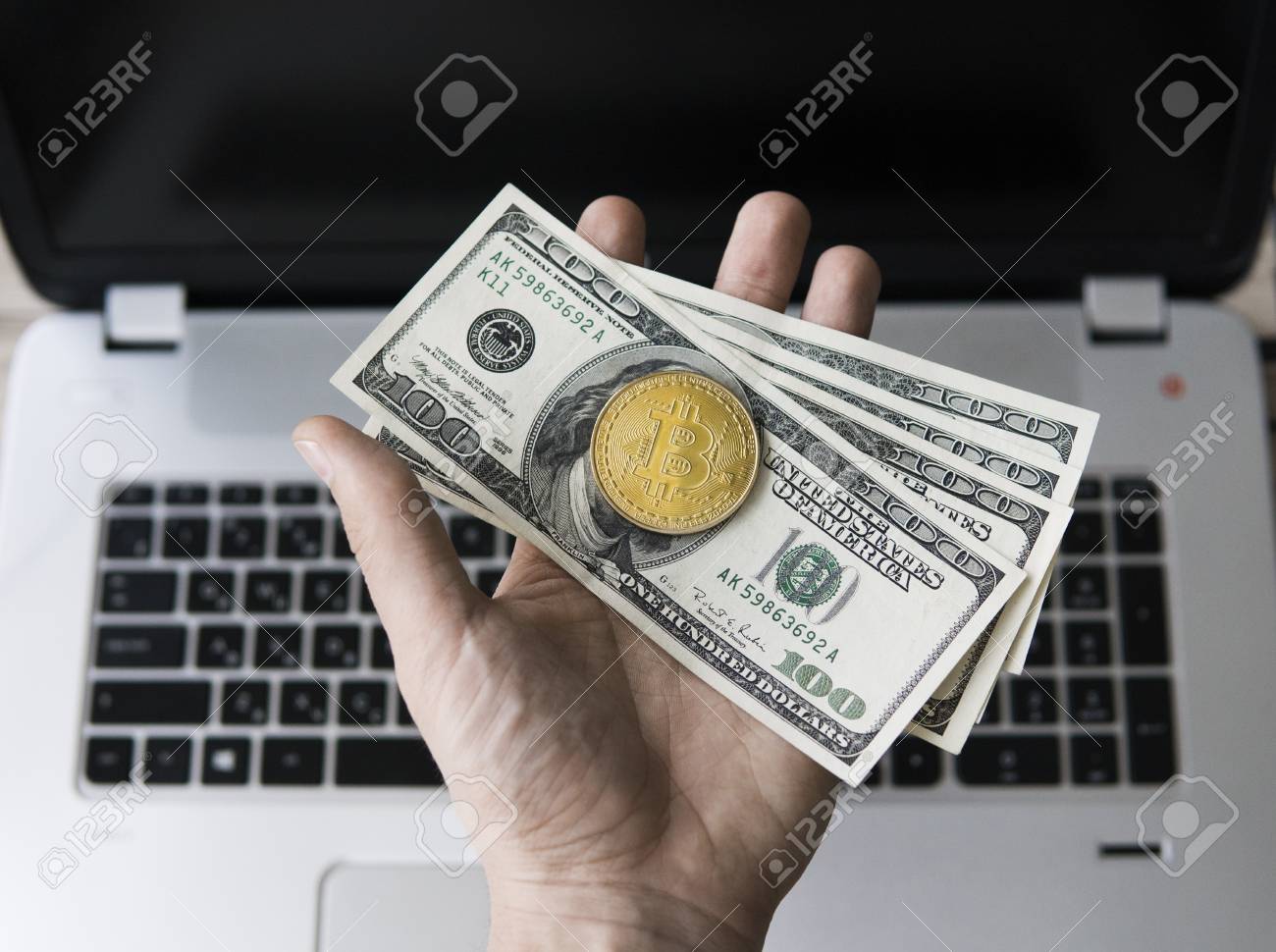 Mano de hombre sosteniendo una moneda de oro de bitcoin con un billete de  un dólar estadounidense contra una computadora portátil en el fondo. Bitcoin  es una moneda criptográfica y un sistema