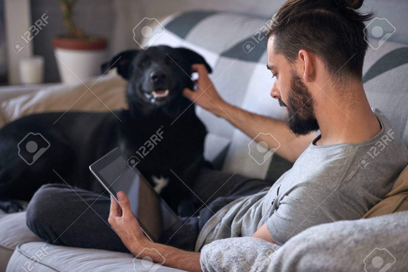 Dog owner petting and scratching his pet on the couch sofa, while surfing the internet on his tablet device, a loving affectionate relationship - 54381115 Dog owner petting and scratching his pet on the couch sofa, while surfing the internet on his tablet device, a loving affectionate relationship - 54381115