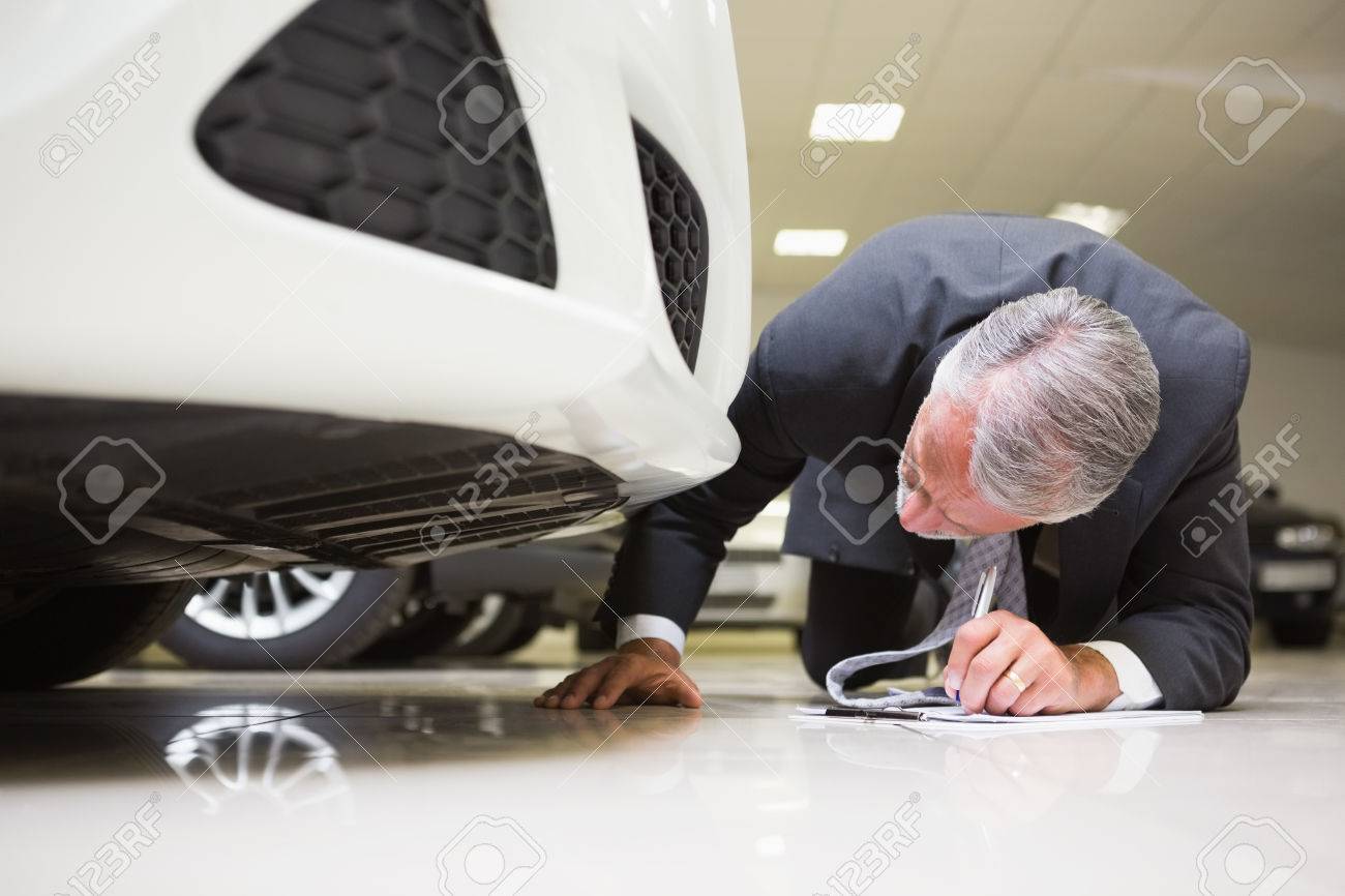36414272-businessman-looking-under-the-car-while-writing-on-clipboard-at-new-car-showroom.jpg