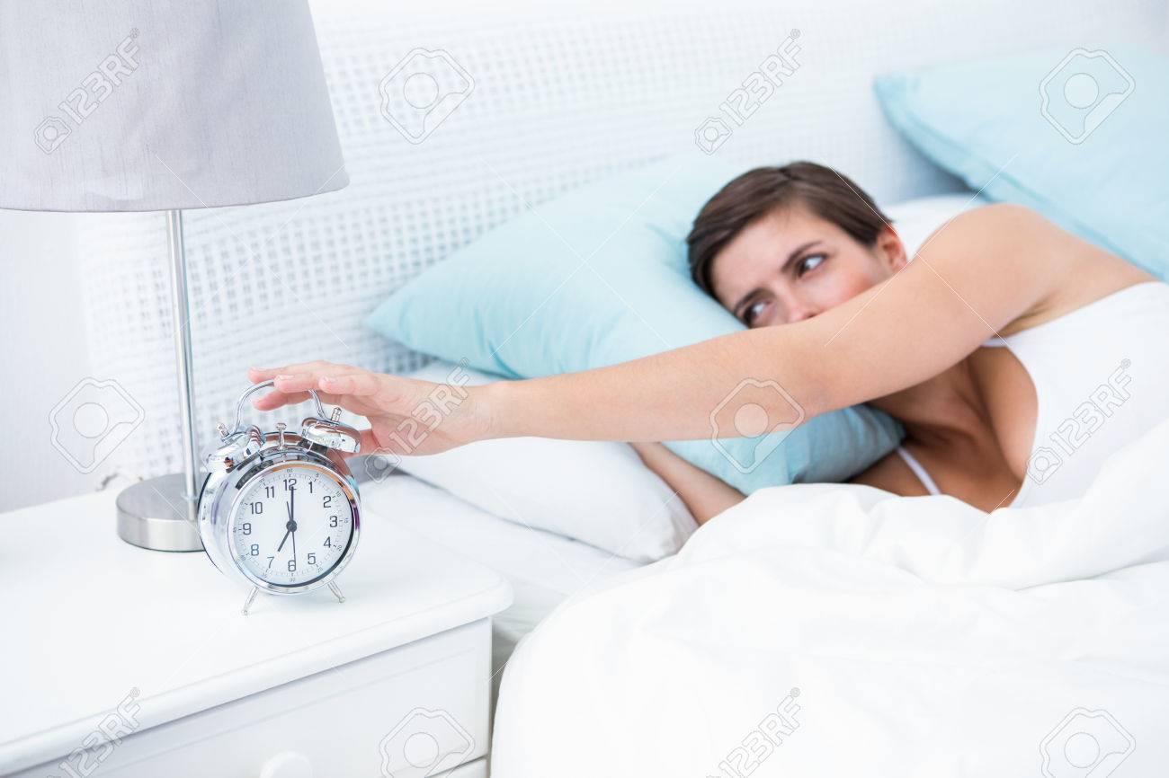 Pretty Brunette Extending Hand To Alarm Clock At Home In The