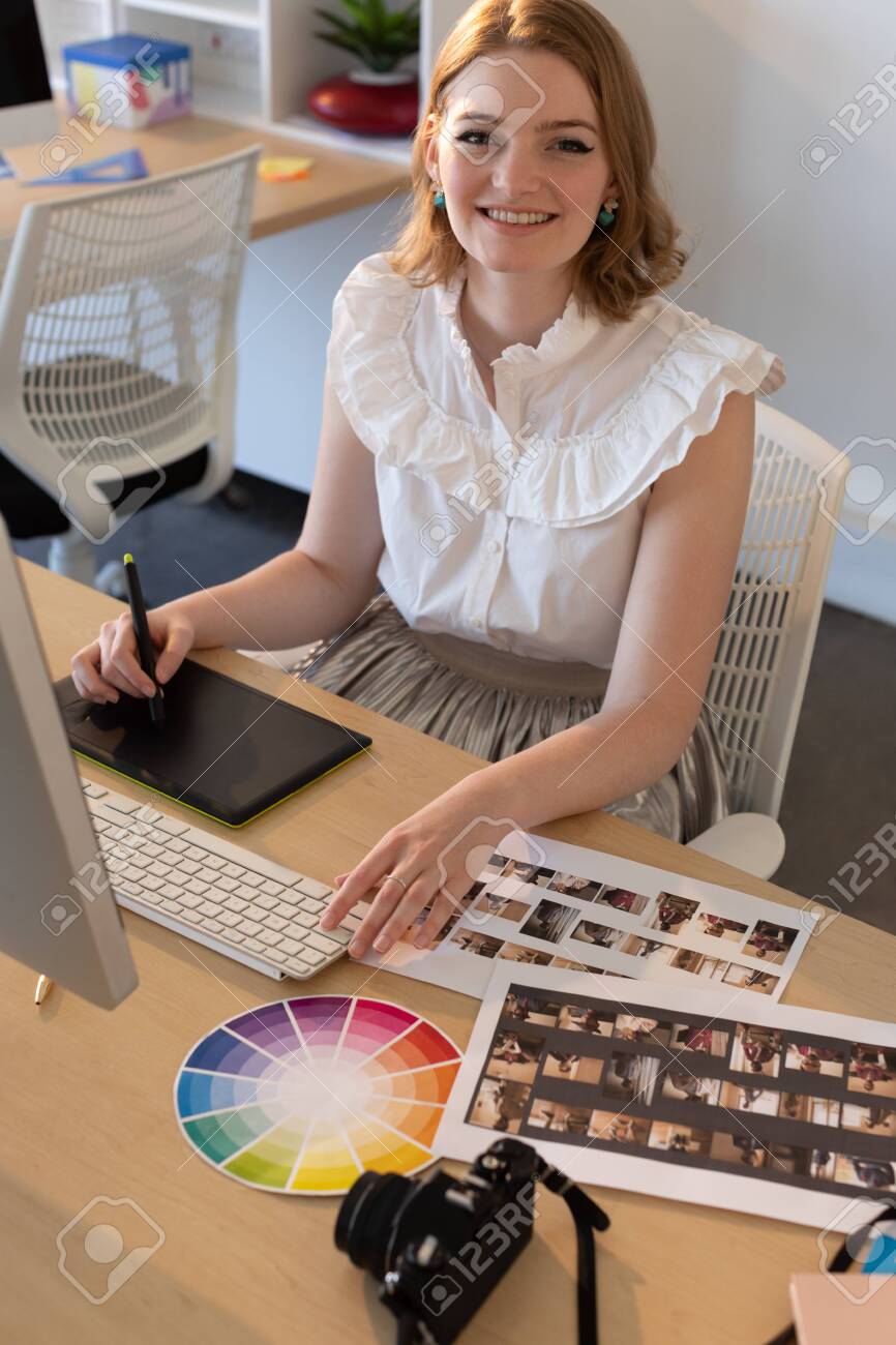 Portrait of young Caucasian female graphic designer working on graphics tablet and computer at desk in the office. She is smiling and looking at camera - 121754173 Portrait of young Caucasian female graphic designer working on graphics tablet and computer at desk in the office. She is smiling and looking at camera - 121754173