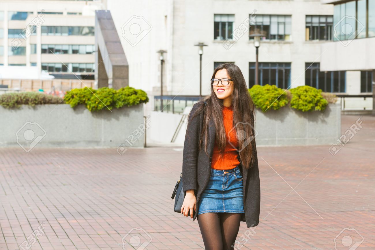美しい中国の女性は 日当たりの良い春の日にロンドンで歩きます デニム スカートと眼鏡とスマート カジュアルな服を着ています 彼女は笑顔で カメラから目を の写真素材 画像素材 Image