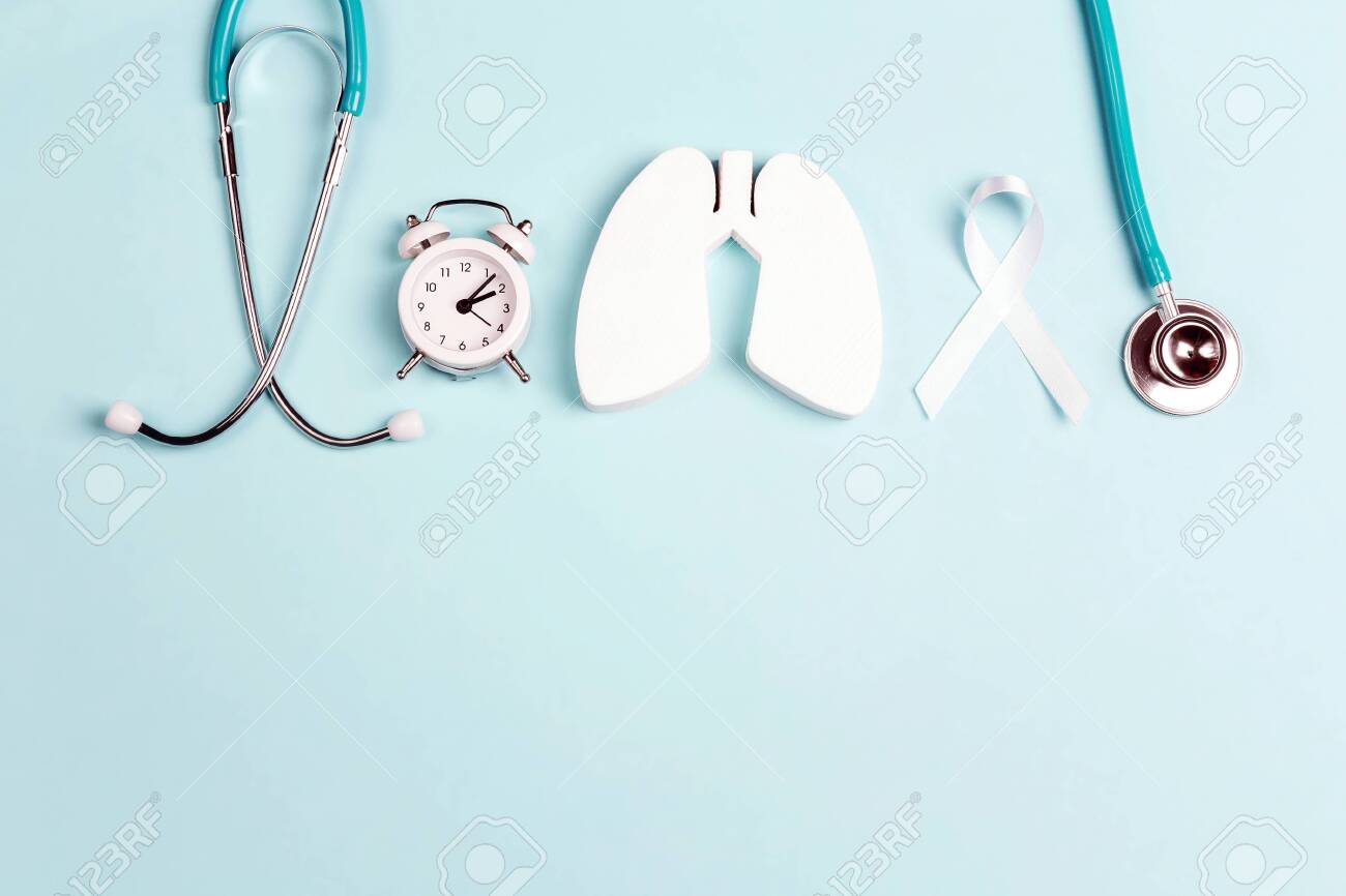 Lung cancer awareness background with white ribbon, alarm clock and stethoscope on blue. Copy space for text. - 131153534 Lung cancer awareness background with white ribbon, alarm clock and stethoscope on blue. Copy space for text. - 131153534