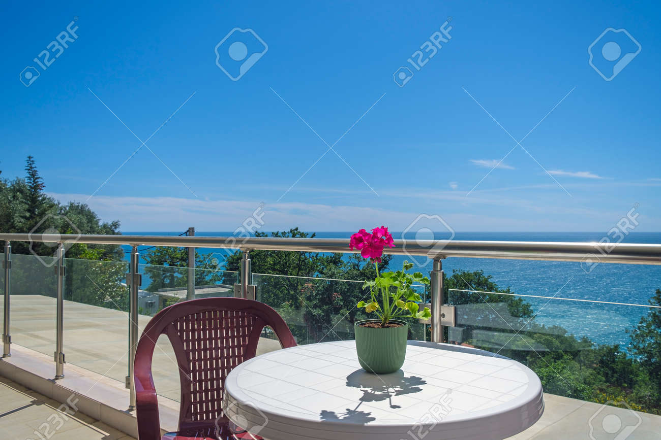 Spacious terrace in the private villa with sea view. The terrace offers view on the sea and coastline, covered by greenery. On the terrace there is a plastic table with a chair, on the table there is a red flower for decoration - 170377744 Spacious terrace in the private villa with sea view. The terrace offers view on the sea and coastline, covered by greenery. On the terrace there is a plastic table with a chair, on the table there is a red flower for decoration - 170377744