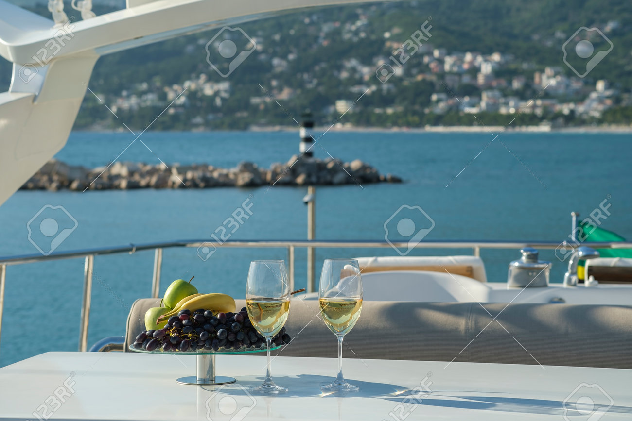Yacht cruise. A table is set on the deck, there are glasses of white wine and a dish of fruit on the table. The yacht offers a view of the lighthouse and the mountains - 170815744 Yacht cruise. A table is set on the deck, there are glasses of white wine and a dish of fruit on the table. The yacht offers a view of the lighthouse and the mountains - 170815744