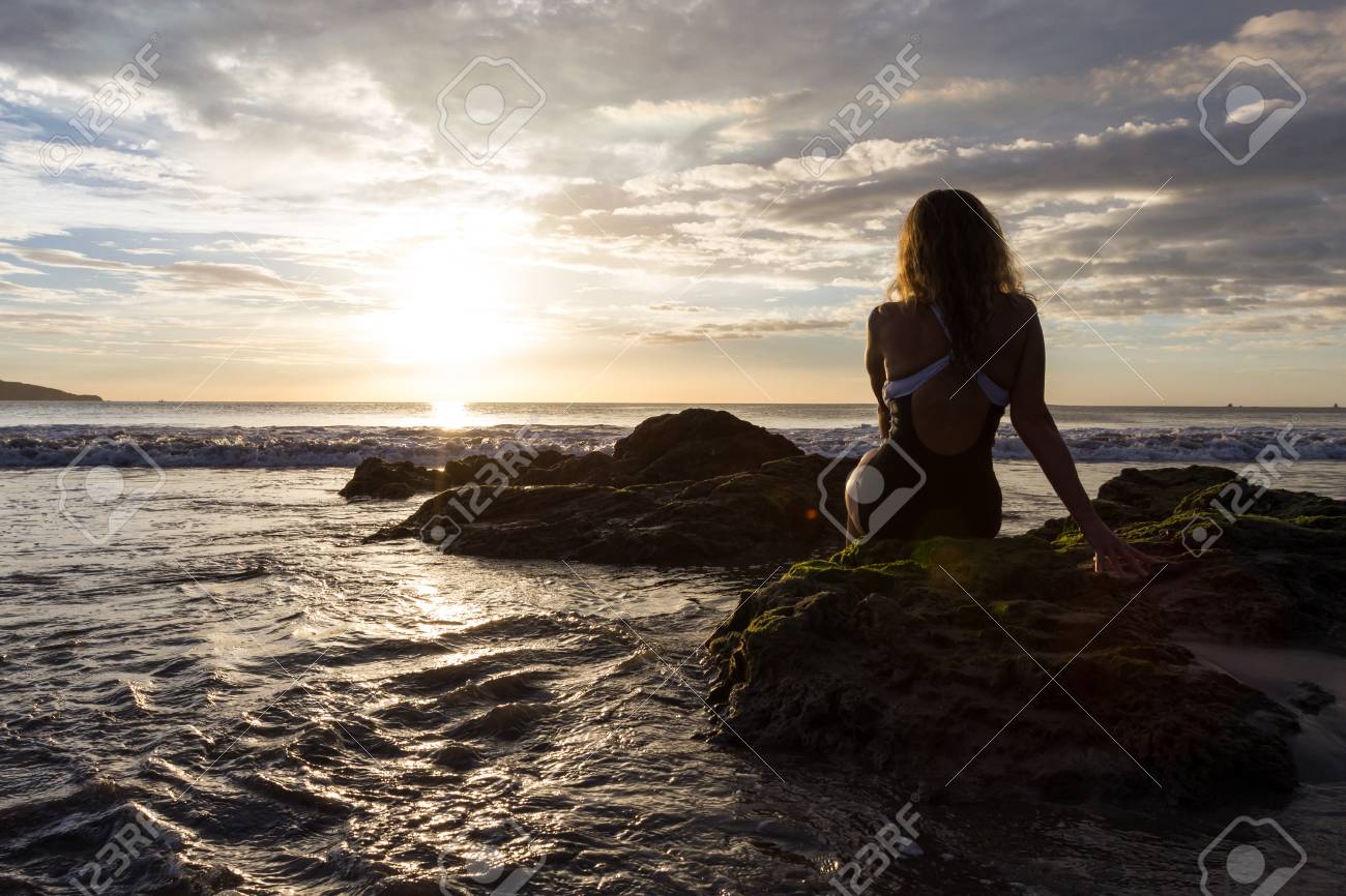 Attractive Woman Sitting On The Rocks Enjoying A Beautiful Sunset