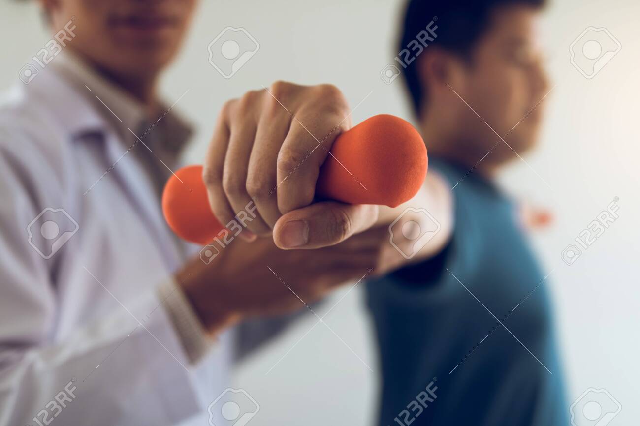 Asian young male physiotherapist helping patient with lifting dumbbells exercises in office. - 123934610 Asian young male physiotherapist helping patient with lifting dumbbells exercises in office. - 123934610
