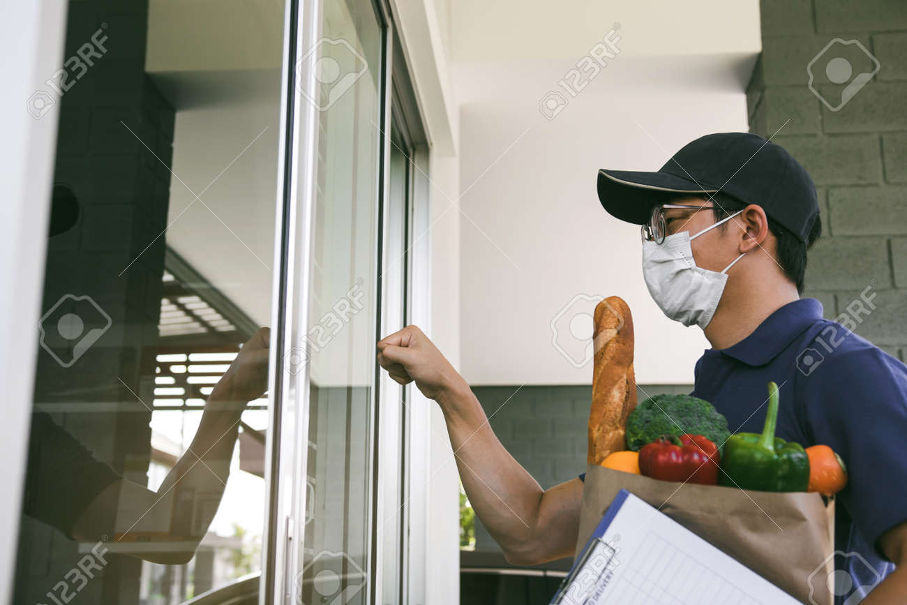 Asian man wearing a mask while holding a food bag standing knocking at the front door. - 152651168 Asian man wearing a mask while holding a food bag standing knocking at the front door. - 152651168