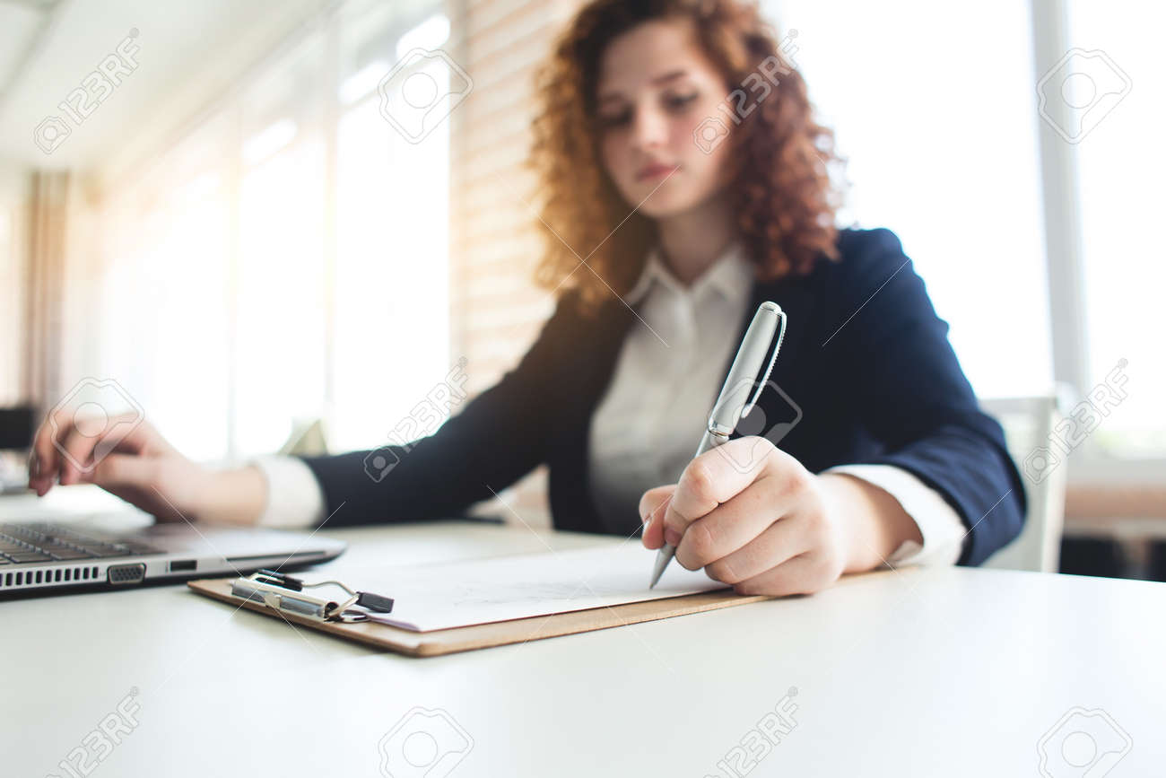 Portret of a successful young curly red-haired woman in a business suit and glasses works in an office on a laptop - 171606168 Portret of a successful young curly red-haired woman in a business suit and glasses works in an office on a laptop - 171606168
