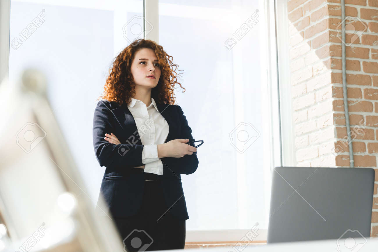 Portrait of a beautiful successful young business woman with red curly hair in the office interior stands near the window - 171943482 Portrait of a beautiful successful young business woman with red curly hair in the office interior stands near the window - 171943482