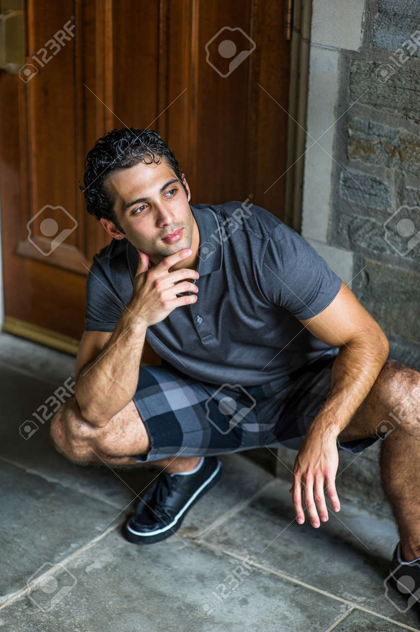 Dressing in a gray polo shirt, pattern shorts and black leather sneakers, one hand touching his chin, a young strong handsome guy is squatting in the front of the door, relaxing and thinking. - 170751271 Dressing in a gray polo shirt, pattern shorts and black leather sneakers, one hand touching his chin, a young strong handsome guy is squatting in the front of the door, relaxing and thinking. - 170751271