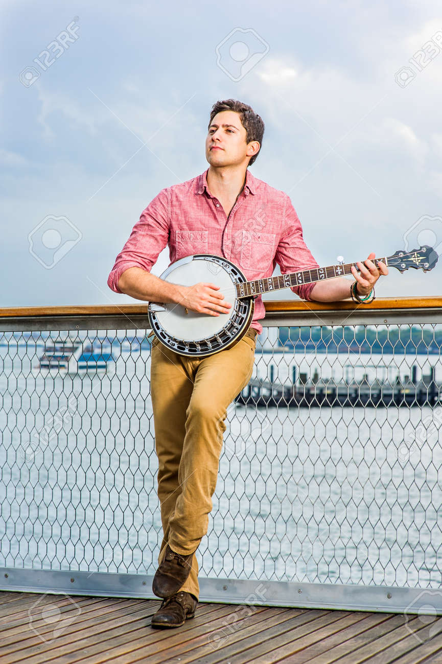 Dressing in light red shirt, dark yellow trousers and brown shoes, looking up and stepping his foot, a young musician is playing a banjo on the deck - 171462667 Dressing in light red shirt, dark yellow trousers and brown shoes, looking up and stepping his foot, a young musician is playing a banjo on the deck - 171462667