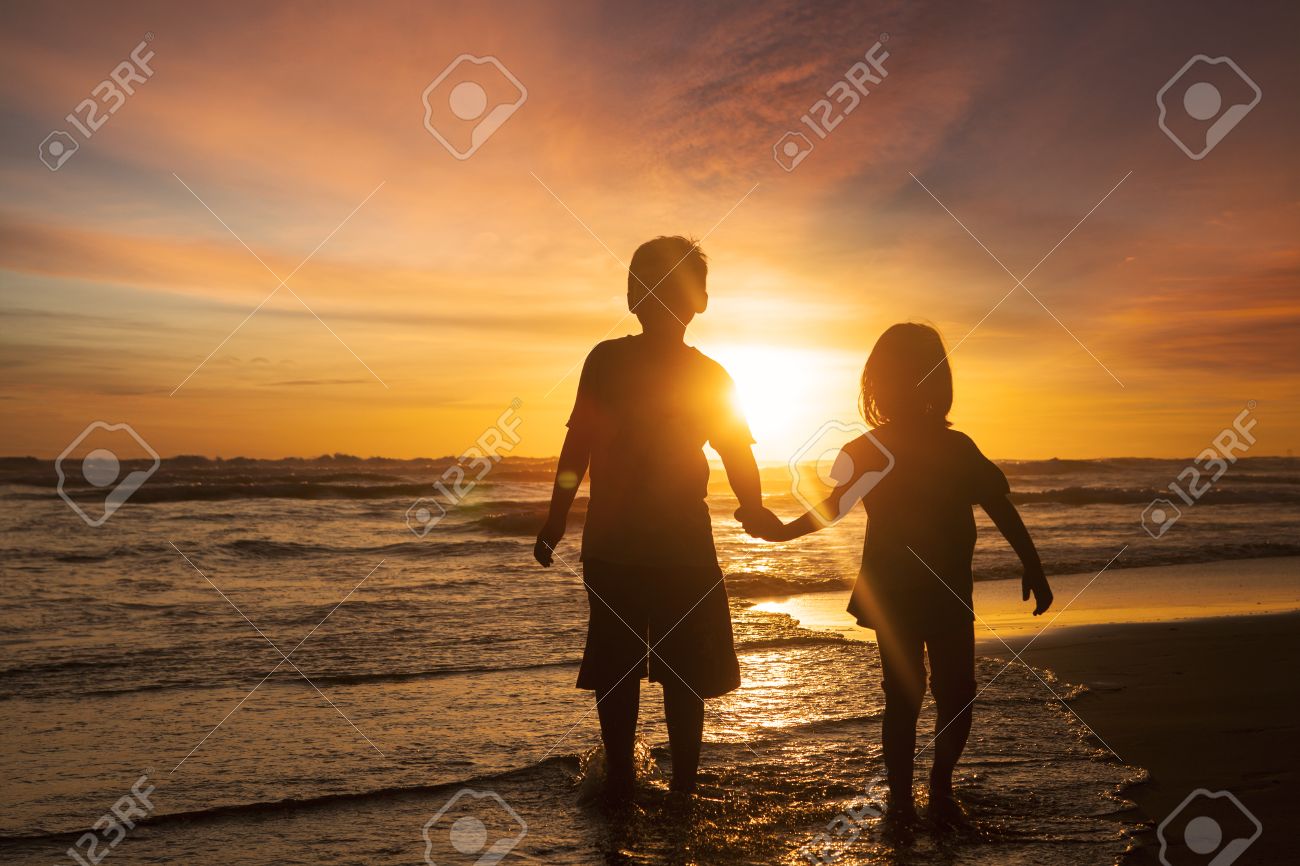 背面に夕日を背景と手を保持しながら ビーチの上を歩いて 2 人の子供のシルエット の写真素材 画像素材 Image