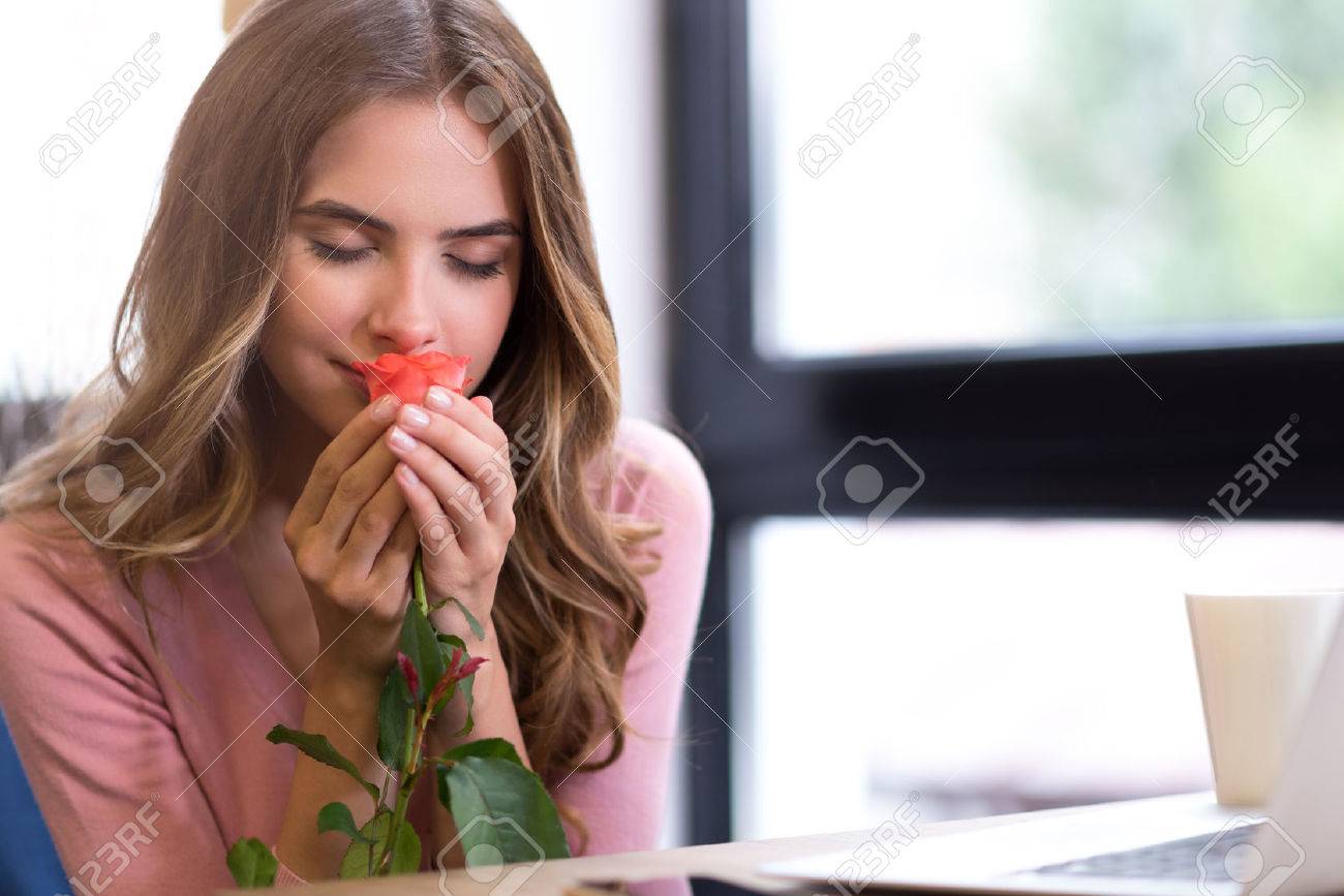 Nice scent. Delighted beautiful young woman sitting at the table and holding rose while smelling it - 61352044 Nice scent. Delighted beautiful young woman sitting at the table and holding rose while smelling it - 61352044