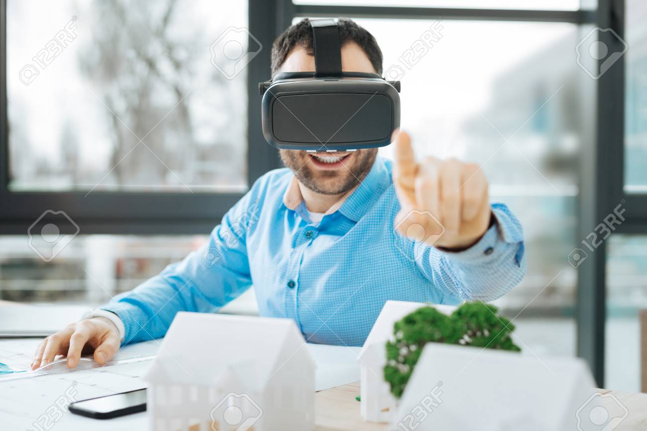 Palpable reality. Handsome young architect wearing a VR headset and raising his finger as if to touch an object in a virtual reality while sitting at his work desk in the office - 93948465 Palpable reality. Handsome young architect wearing a VR headset and raising his finger as if to touch an object in a virtual reality while sitting at his work desk in the office - 93948465
