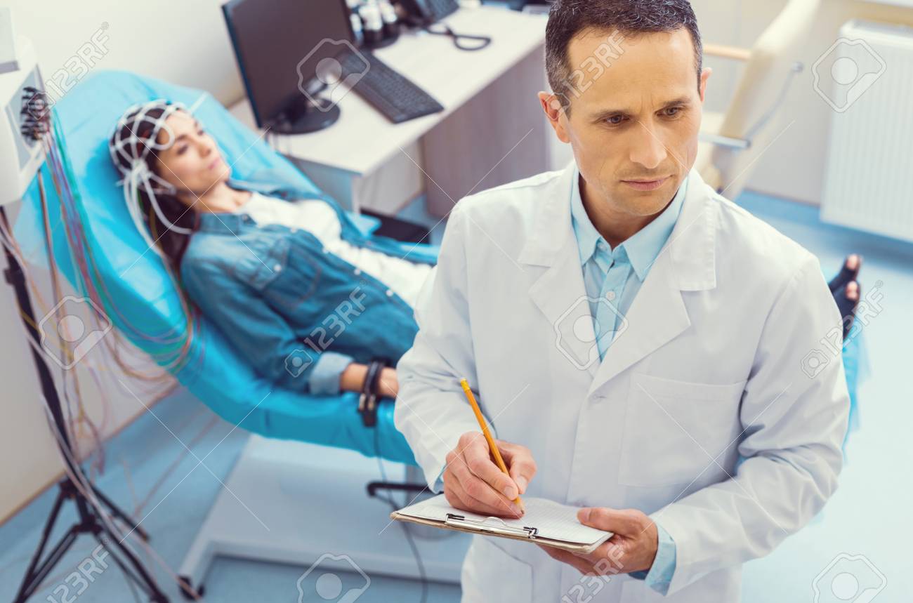 Making a diagnosis. Top view on a pensive mature medical worker holding a clipboard while writing something down during an electroencephalography analysis. - 107807217 Making a diagnosis. Top view on a pensive mature medical worker holding a clipboard while writing something down during an electroencephalography analysis. - 107807217