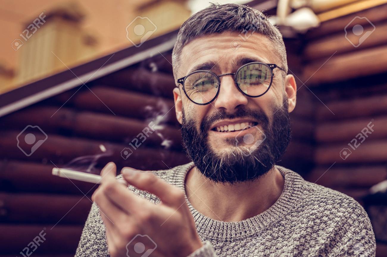 Unhealthy Lifestyle. Joyful Good Looking Man Smiling To You While Enjoying  Smoking Cigarettes Фотография, картинки, изображения и сток-фотография без  роялти. Image 117796974
