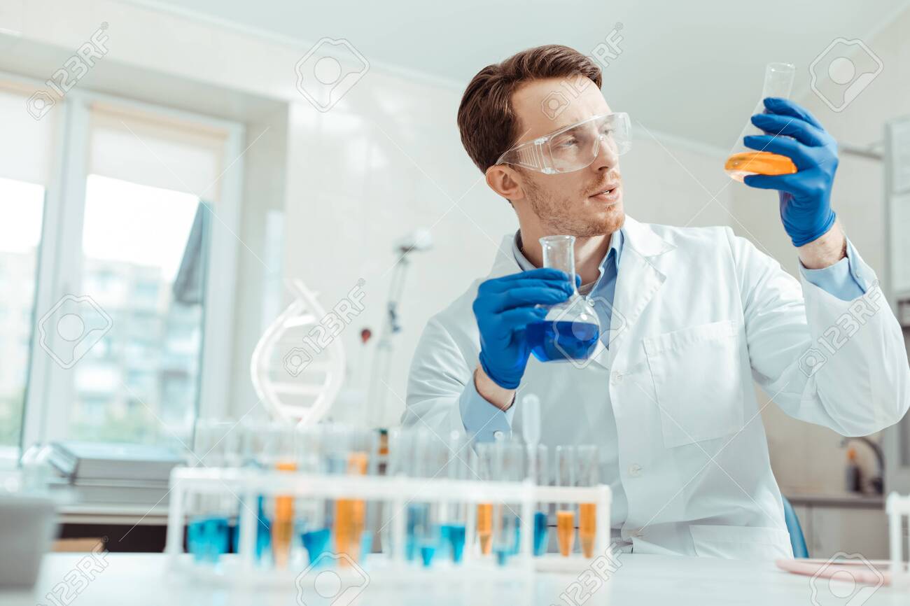 Professional chemist. Nice handsome man holding two different flasks while working in the chemical lab - 127134357 Professional chemist. Nice handsome man holding two different flasks while working in the chemical lab - 127134357